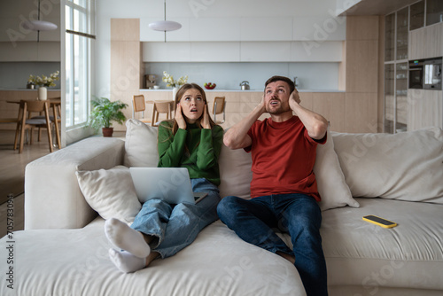 Annoyed unhappy couple sitting on sofa with vexed face expression, looking upward, closing ears because of loud sound, noise from neighboring flat. Emotional stress, discomfort caused by neighborhood