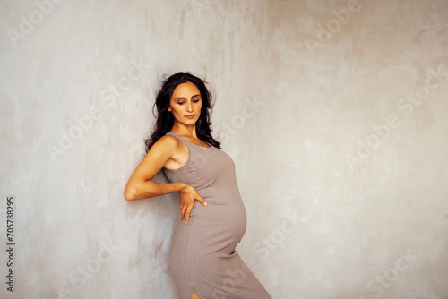 A portrat of beautiful pregnant young woman. Brunette female in a beige dress stands against a light wall. Studio shot of a woman who is expecting a baby. photo