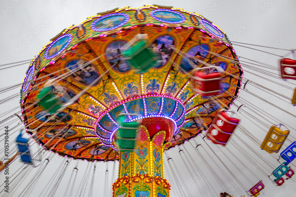Um brinquedo de rotação, grande e colorido, no parque de diversões com poucas pessoas, em um dia nublado. Foto feita de baixo para cima. - obrazy, fototapety, plakaty 