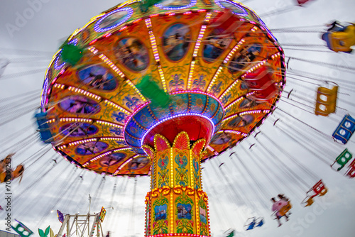 Um brinquedo gigante de rotação, com várias cadeiras no alto, borradas pela velocidade do movimento, no parque de diversões com poucas pessoas, em um dia nublado. Foto feita de baixo para cima.