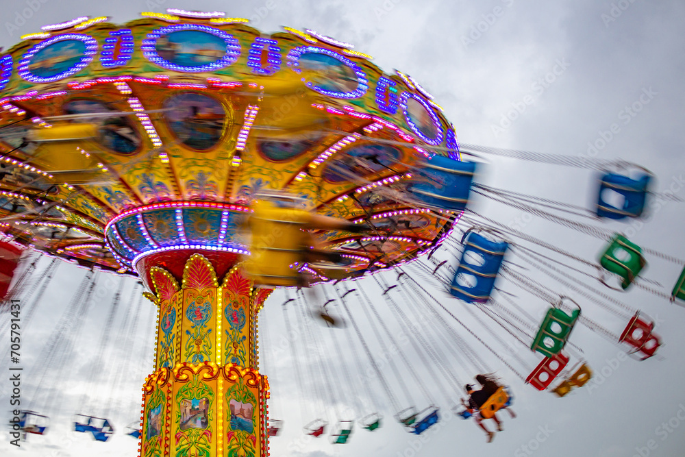 Um brinquedo gigante de rotação, com várias cadeiras no alto, borradas pela velocidade do movimento, no parque de diversões com poucas pessoas, em um dia nublado. Foto feita de baixo para cima. - obrazy, fototapety, plakaty 