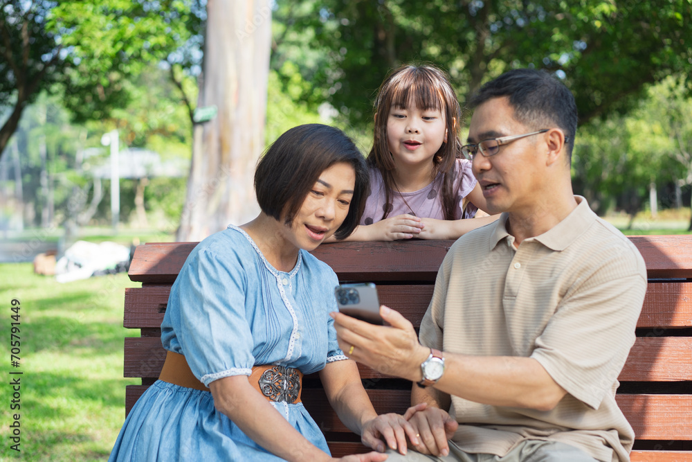 Happy asian family of three having fun watching a phone together in the summer outdoors at the park.