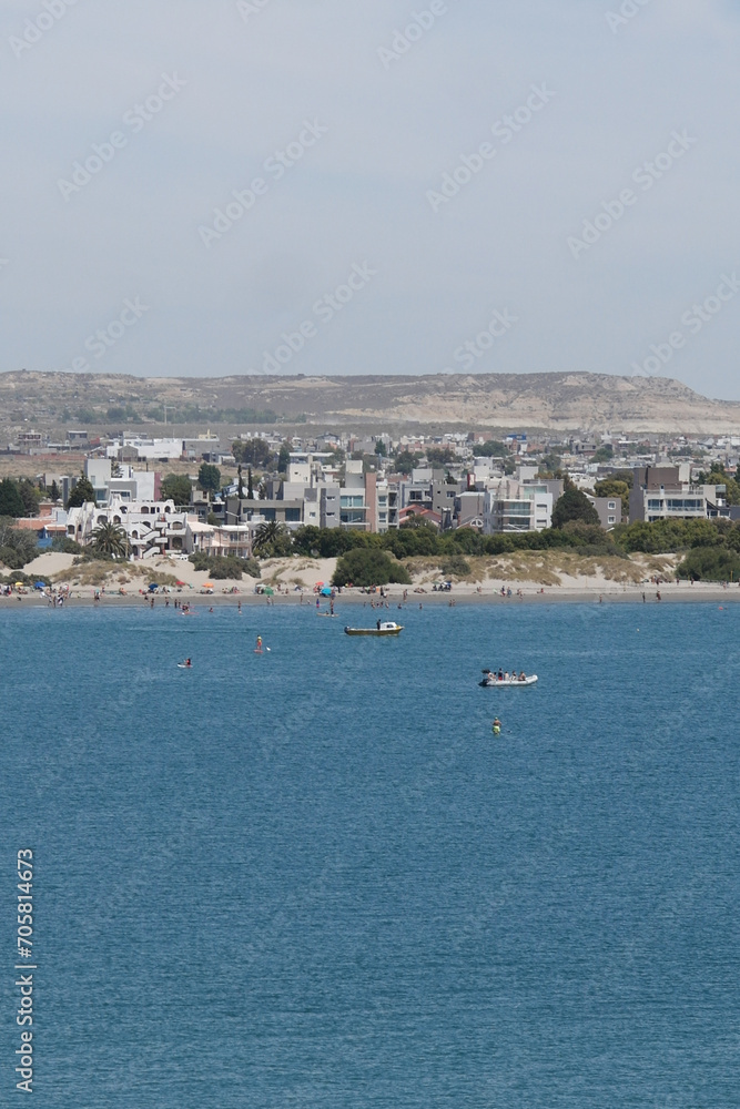 The image shows a coastal town with buildings near the shore, a boat on the water, and hills in the background. The main focus is on the serene coastal town with white buildings clustered near the sho