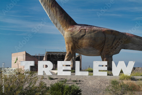 The image shows a billboard that advertises the presence of the world’s largest dinosaur in Trelew. The main focus is a large white billboard with black and blue text, set against a clear sky
