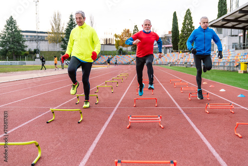 Three unrecognizable senior runners on a track doing high knee drills over yellow and red hurdles for agility