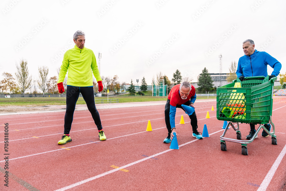Obraz premium Three senior male runners on a track, one standing, one arranging cones, and another with a cart of equipment