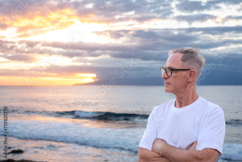 Senior mature man at the sea beach at sunset light looking the horizon over sea admiring the orange sunray and cloudy sky, handsome man enjoys a relaxed retirement lifestyle