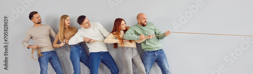Group of happy adult friends playing tug of war and applying teamwork and unity strategy. Team of cheerful confident people standing together by grey wall pulling rope in one direction. Header, banner photo