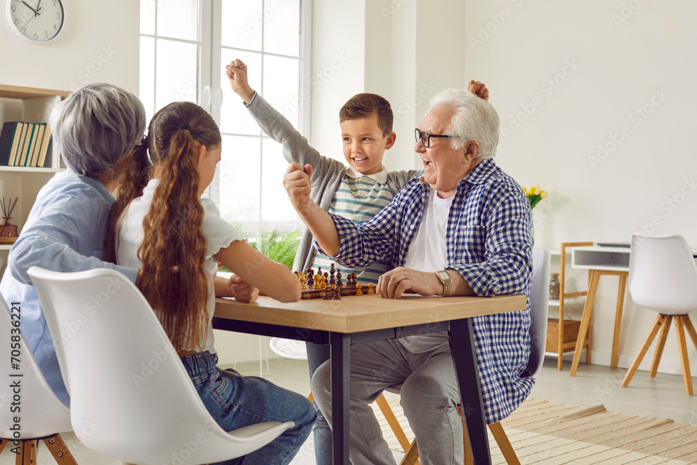Children and grandparents play chess together. Happy boy and grandfather win, celebrate victory, shout yes hooray and do fist up gestures while sitting at table with sad grandma and granddaughter - obrazy, fototapety, plakaty 