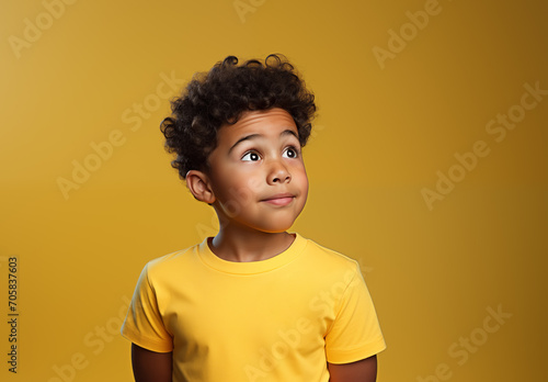 Young mixed race boy with curly hair in a yellow T-shirt looking up sideways with a curious expression on his face. Dark-skinned child isolated on yellow background with copy space