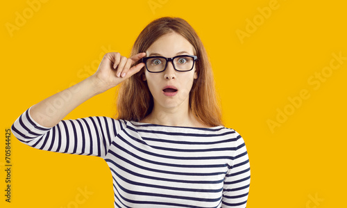 Young student girl very surprised by something. Beautiful nerdy young woman in striped top and glasses isolated on yellow background looking at camera with funny, shocked, astonished face expression photo