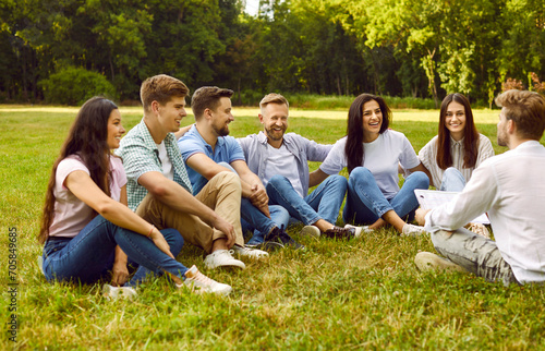 Man taking interview from a group of a young smiling happy people students sitting on a green grass in the summer park and answering on social survey or having a training outdoors.