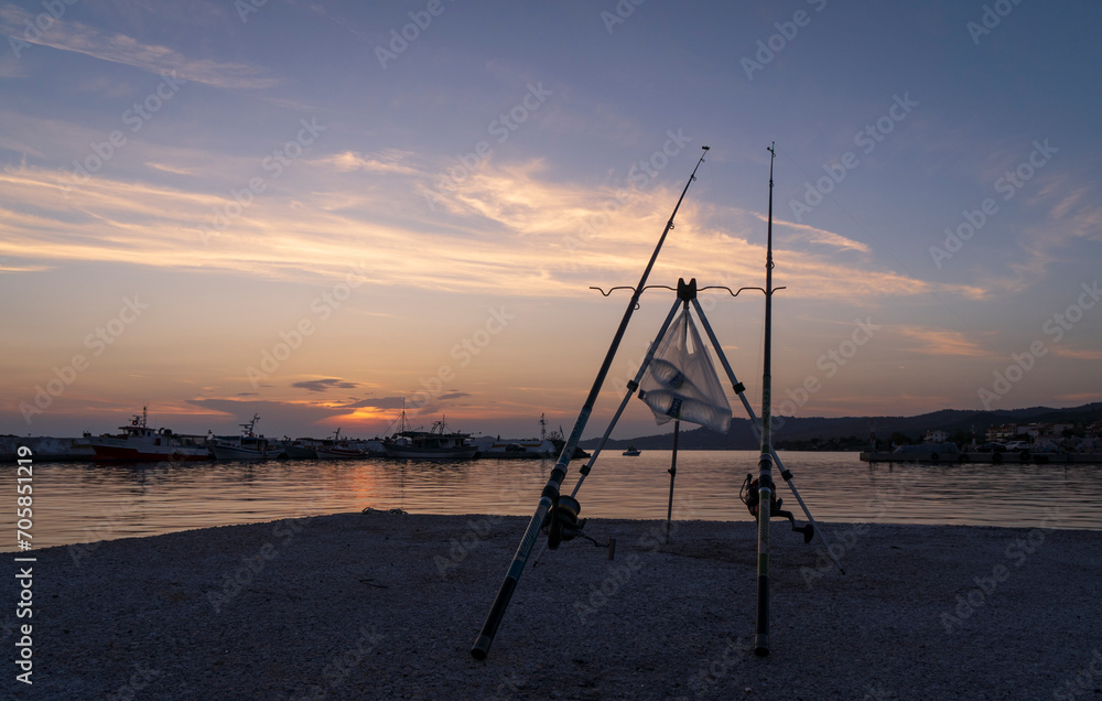 Fishing rods stand on the edge of a pier in a bay with fishing boats at sunset in Skioni, Greece Fishing rods stand on the edge of a pier in a bay with fishing boats at sunset in Skioni, Greece