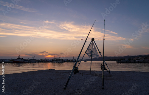 Fishing rods stand on the edge of a pier in a bay with fishing boats at sunset in Skioni, Greece