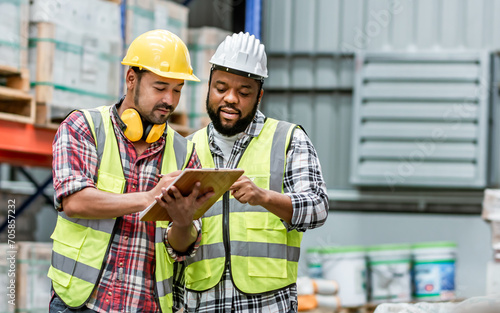 Two diverse professional male workers holding board, checking shipping stocks in storage, warehouse or factory for delivery, wearing safety hat. Diversity, Commercial Industry Business Concept.