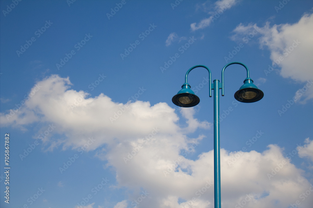 Teal blue metal street lamp stands against fluffy white clouds and bright blue sky. Teal blue metal street lamp stands against fluffy white clouds and bright blue sky.