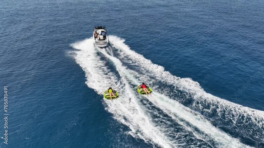 Aerial drone video of extreme inflatable towable tube cruising in high speed attached on powerboat with children on sofa ride, water sports in tropical exotic bay with deep blue crystal clear sea Aerial drone video of extreme inflatable towable tube cruising in high speed attached on powerboat with children on sofa ride, water sports in tropical exotic bay with deep blue crystal clear sea