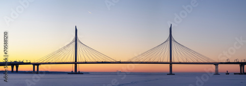 Panoramic view: Large cable-stayed expressway bridge at sunset over an ice-covered bay with an airplane flying high