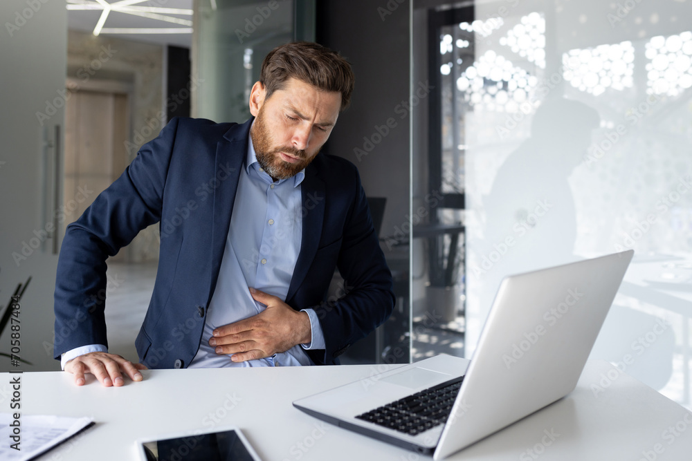 A young male businessman is sitting in the office at a desk with a laptop and holds his stomach with his hands twisted. Feels pain and discomfort A young male businessman is sitting in the office at a desk with a laptop and holds his stomach with his hands twisted. Feels pain and discomfort