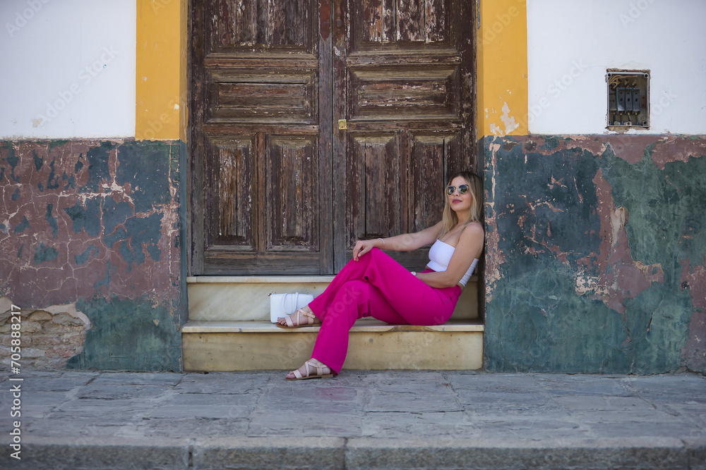 South American woman, young, beautiful and blonde with white top, pink pants and sunglasses, posing sitting on the windowsill of a wooden door, relaxed and quiet. Concept trend, beauty, fashion.