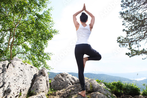 Attractive young woman doing a yoga pose for balance and stretching staying on top of high rock in the mountains