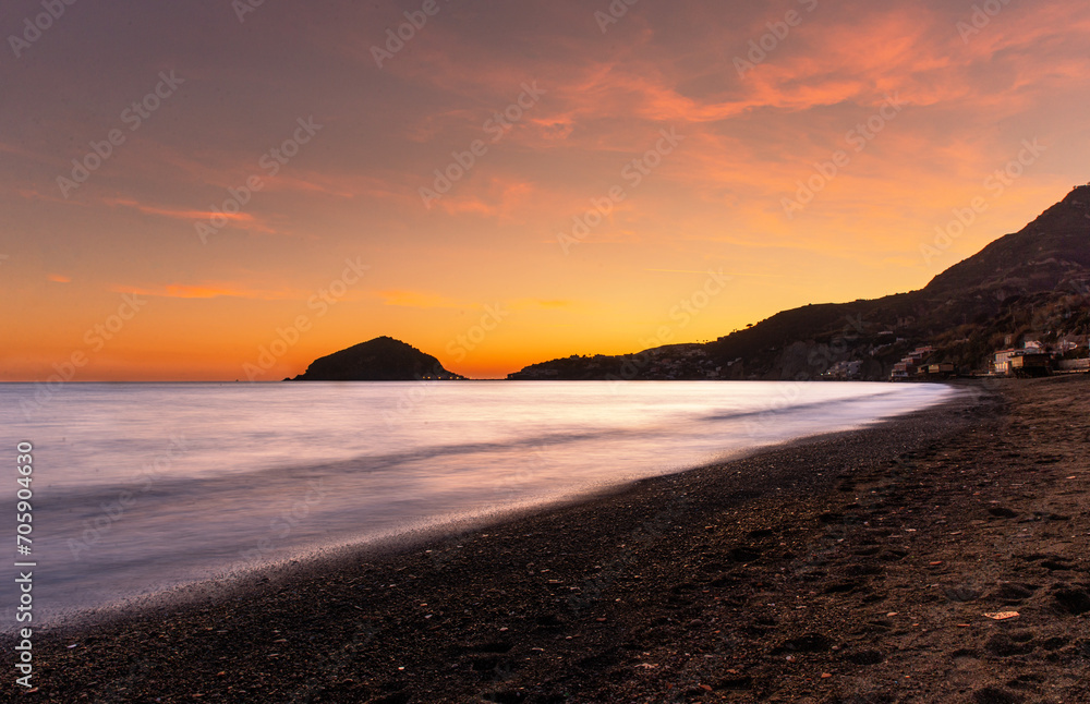 tramonto visto da una delle spiagge dell'isola d'Ischia, acqua con effetto seta illuminata dai raggi del sole durante il tramonto, colori caldi del tramonto, scogli e montagne in penombra