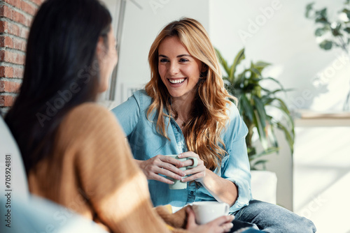 Two smiling young women talking while drinking coffee sitting on couch in the living room at home. photo