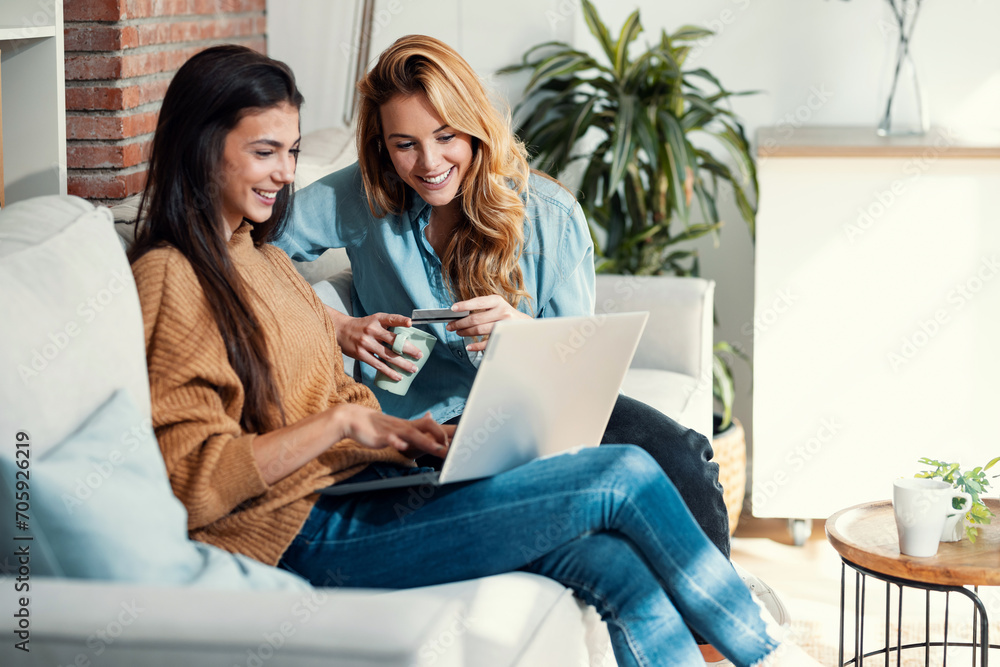 Two happy young women using computer while paying with credit card their vacation sitting on the couch at home.