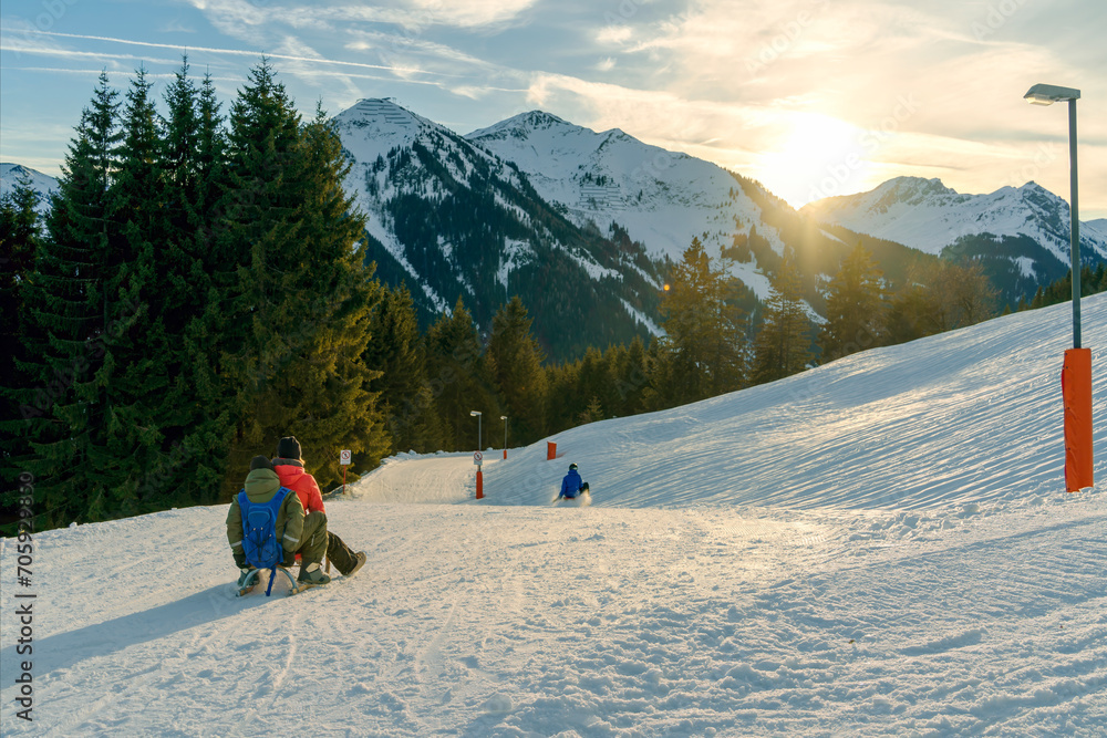 Family moments as they sled down a sun-kissed snowy hill, with the majestic Alps in the background, embodying the joy of winter leisure