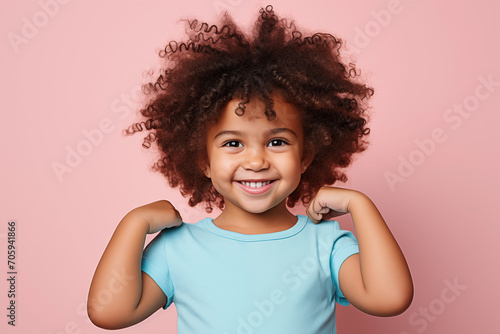 Cute little African American child with curly afro hair in blue t-shirt smiling and showing a gesture of strength on pink background with copy space