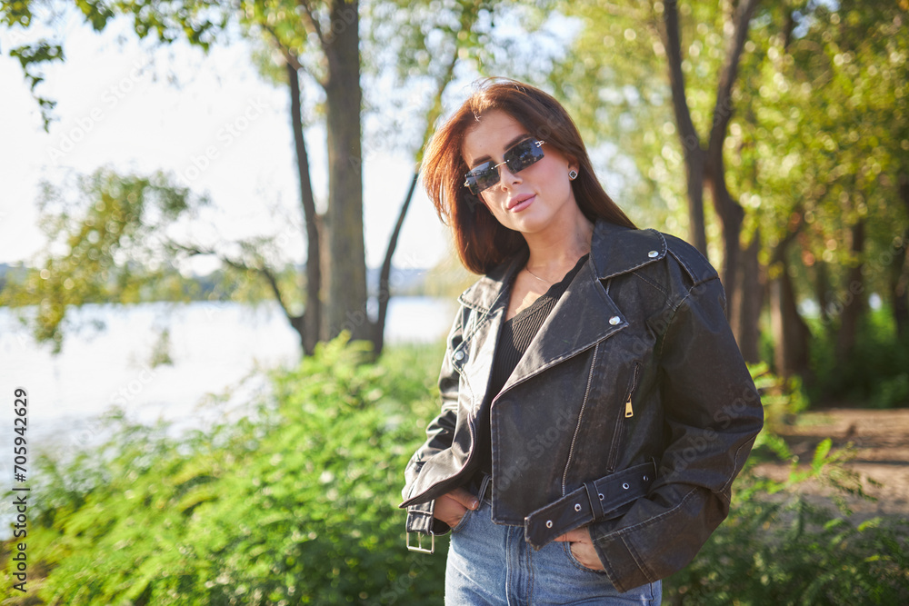 A young brunette, adorned in a leather jacket, poses against the backdrop of nature A young brunette, adorned in a leather jacket, poses against the backdrop of nature