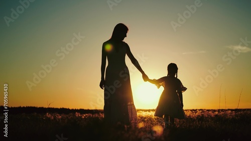 Silhouettes of mamma holding hand of daughter and talking about childhood while walking with child in sunlit field. Silhouettes of mamma with daughter sharing secrets at setting sun in park © SUPER FOX