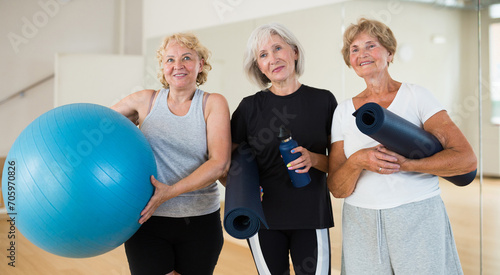 Portrait of three positive mature women standing in a fitness studio after a workout  holding a fitball and twisted sports ..mats in their hands