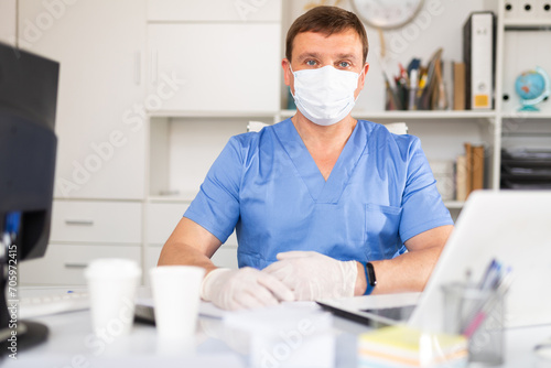 Focused man doctor wearing medical facial mask and gloves working in medical office using laptop computer