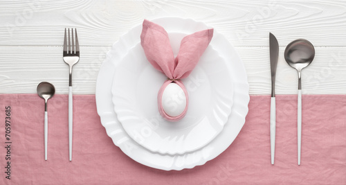 Easter holiday table setting with plate and cutlery, invitation concept. White wooden background. Bunny made of pink linen napkin and egg. Top view. Brunch or dinner.