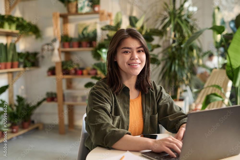 Medium shot of young female plant store owner sitting at her workplace with laptop smiling at camera Medium shot of young female plant store owner sitting at her workplace with laptop smiling at camera