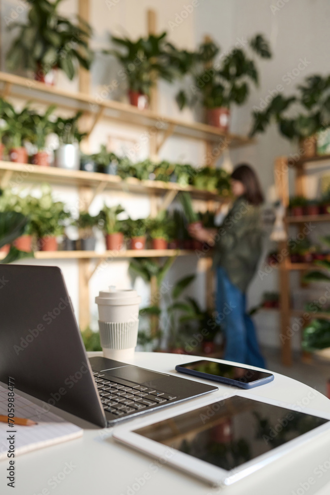 Electronic devices displayed on desk in plant store, worker arranging plants on background Electronic devices displayed on desk in plant store, worker arranging plants on background