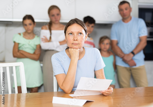 Frustrated woman sits in kitchen and reads unpleasant news in letter. Rent increase, penalty for late payment of goods, business problems. Blurred family husband and children in background