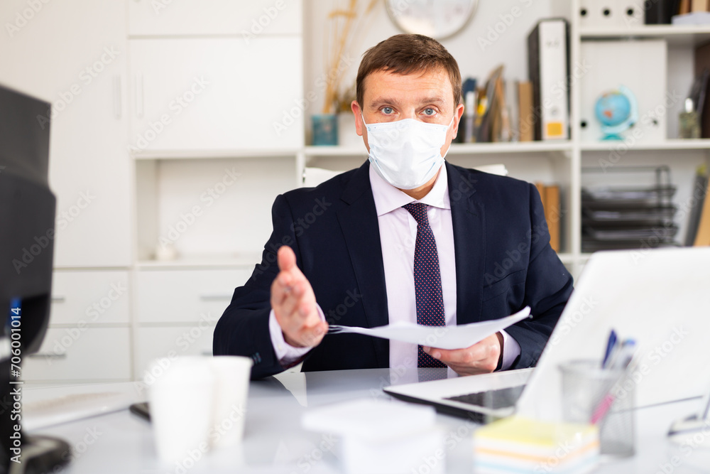 Confident manager in disposable surgical mask discussing financial report or explaining task while sitting in office. Concept of new life reality and precautions in COVID 19 pandemic