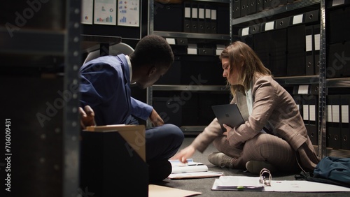 Black policeman and female law officer sitting on office floor with paperwork and laptop as crime investigation is being conducted. Multiracial investigators review and debate case evidence and hints. photo