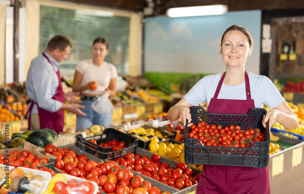 Smiling successful female owner of greengrocery store, preparing fresh fruits and vegetables for sale, placing plastic crates with ripe red cherry tomatoes on counter..