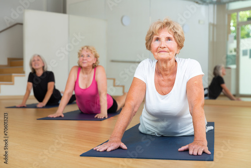 Positive senior lady with group of aged women exercising Hatha yoga poses in modern yoga studio, doing Upward Facing Dog stretching pose.