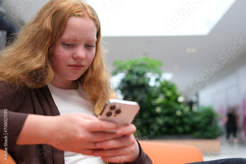 girl in Mall sits using phone to communicate with friends online shopping order on Internet catching Internet games typing text news in background people are walking, unrecognizable, around the store.