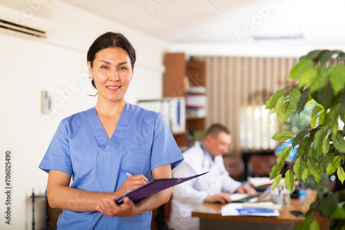 Portrait of polite Asian female doctor wearing blue uniform meeting patient in clinic, filling out medical form at clipboard