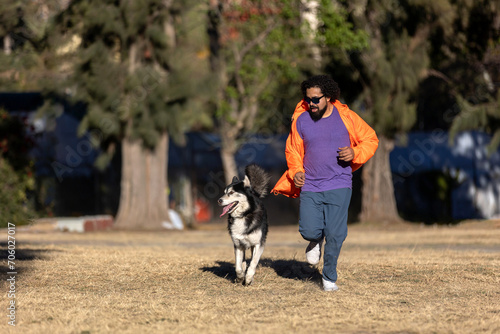 Latin man with beard, curly hair, sunglasses and casual clothes running with his husky dog in a park