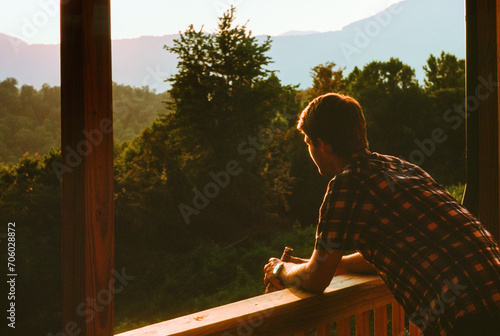 A young man in a flannel shirt leans on the railing of the porch of a rustic cabin overlooking forests and mountains at sunset near Oak Ridge, Tennessee. He holds a glass of beer in his hands. Closeup photo