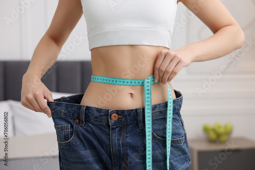 Slim woman wearing big jeans and measuring waist with tape in room, closeup. Weight loss