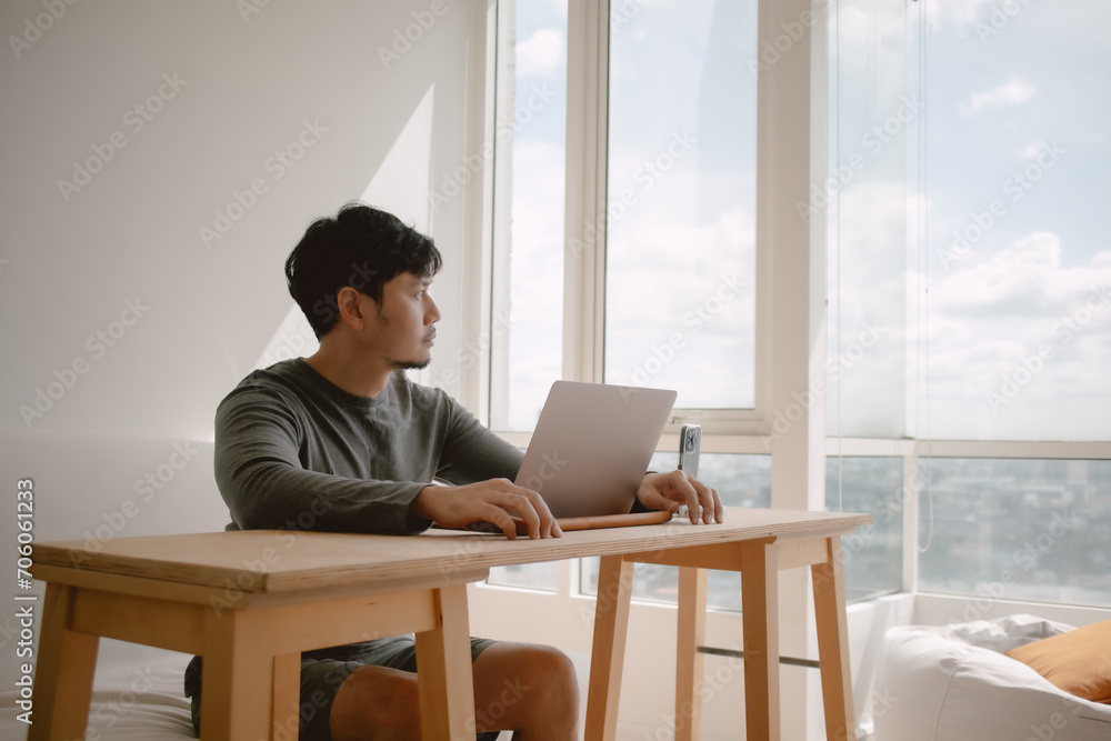Asian man working with computer in apartment with city view at the windows in morning light. Asian man working with computer in apartment with city view at the windows in morning light.