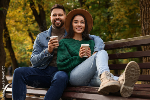 Happy young couple with cups of coffee spending time together on wooden bench in autumn park