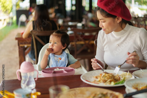 mother with toddler girl eating food and vegetable by self feeding BLW or baby led weaning on highchair in restaurant photo